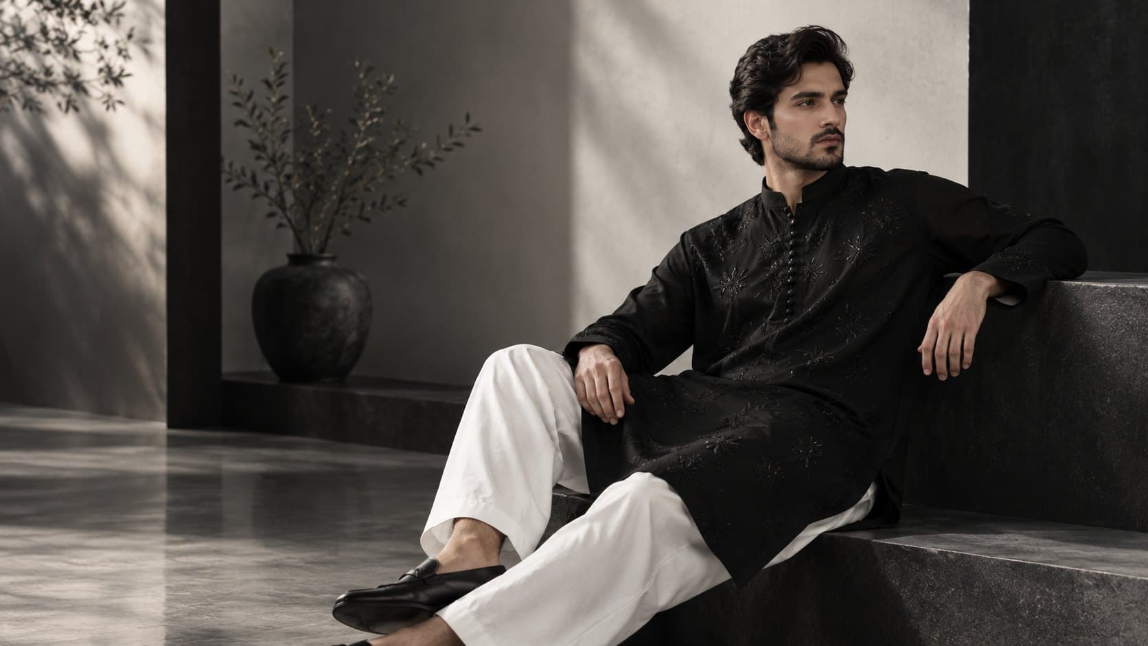 A man in a black kurta with tone-on-tone floral embroidery and white shalwar, seated on a dark stone ledge